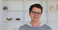 Nahaufnahme einer lächelnden Frau mit kurzen Haaren und Brille vor Regal