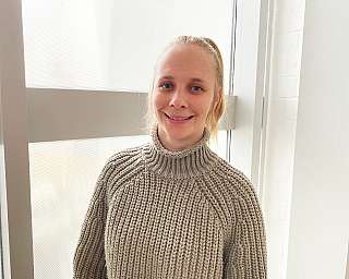 Eine Person in einem grauen Pullover steht vor einem Fenster.