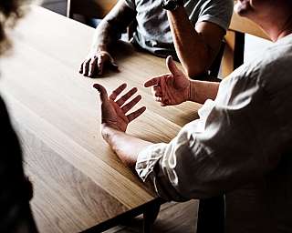 Drei Personen führen ein Gespräch, Hände gestikulieren über einem Holztisch