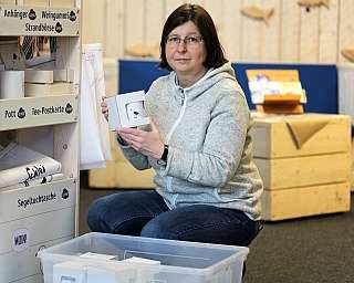 Frau mit Brille hält eine weiße Verpackung mit Tasse, neben Holzhändler-Regal mit Produkten