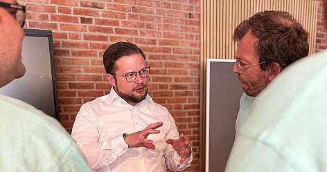 Mann im weißen Hemd spricht mit Personen in hellen T-Shirts vor Backsteinwand