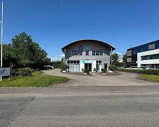Modernes zweistöckiges Bürogebäude mit Arkadeingang und davor parkenden Autos bei klarem Himmel und grüner Umgebung im Sommerlicht, aufgenommen von vorne an der Straße aus betrachtet, mit einem Schild und Flaggenmast links davor, daneben ein zweites Gebäu