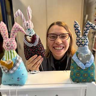 Lächelnde Frau mit Brille hinter Nähmaschine und drei bunten Stoffhasen