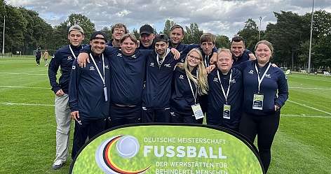 Gruppenfoto unserer Fussball-Mannschaft, die vor einem Schild steht. Auf dem Schild steht: Deutsche Meisterschaft Fussball, der Werkstätten für behinderte Menschen, DFB-Stiftung Sepp Herberger, Deutscher Behindertensportverband e.V. und BAG WfbM